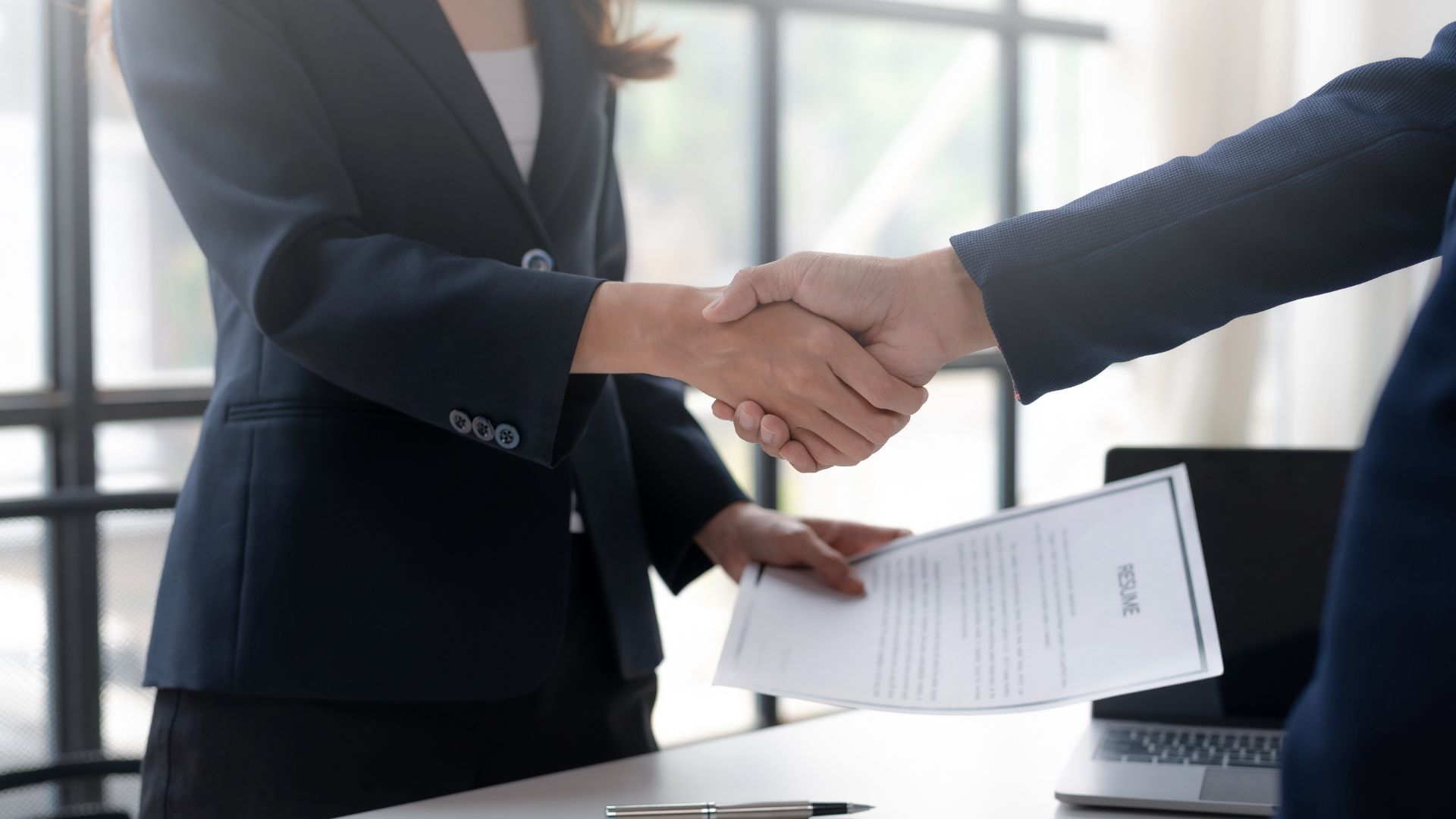 A woman in a business suit shaking hands with a man in a business suit while holding a document in a corporate office setting.