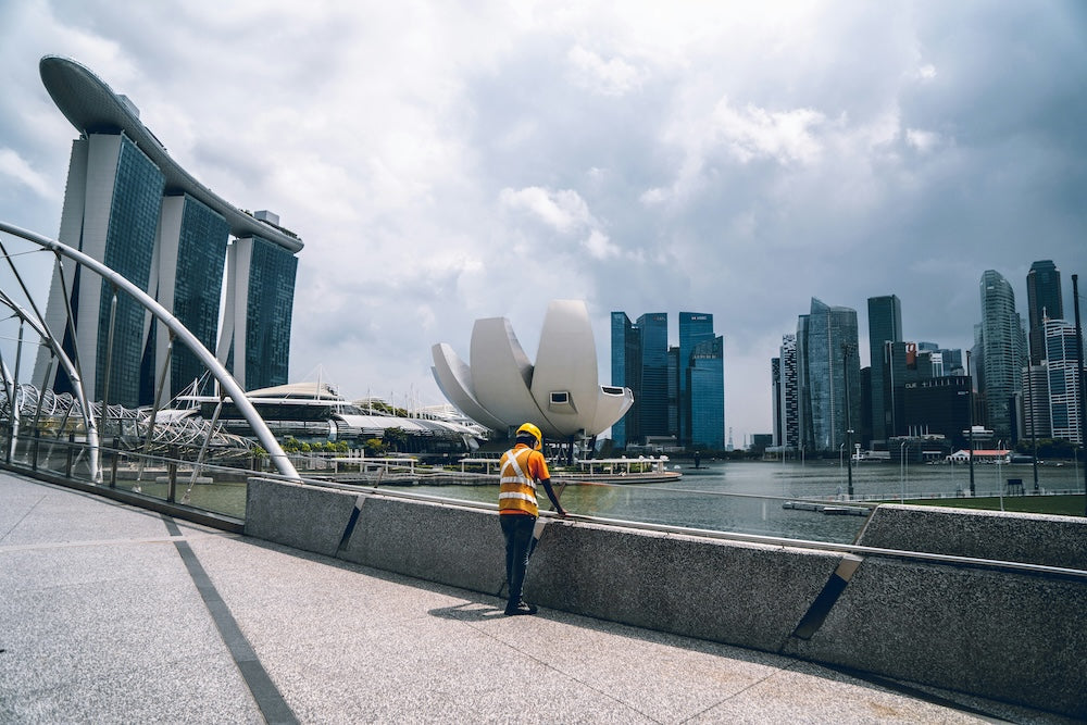 A Bangladeshi construction worker at Esplanade Park overlooking the waterfront, with the ArtScience Museum, Marina Bay Sands, and Singapore’s CBD skyline in the background.
