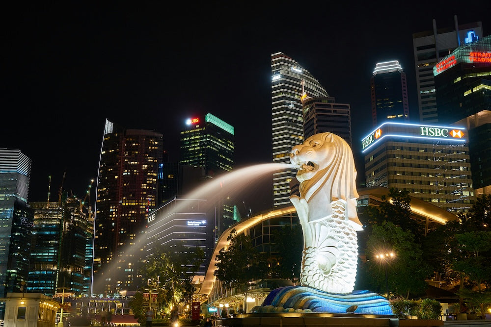 Merlion statue spraying water at night with Singapore skyline and illuminated skyscrapers in the background at Marina Bay.