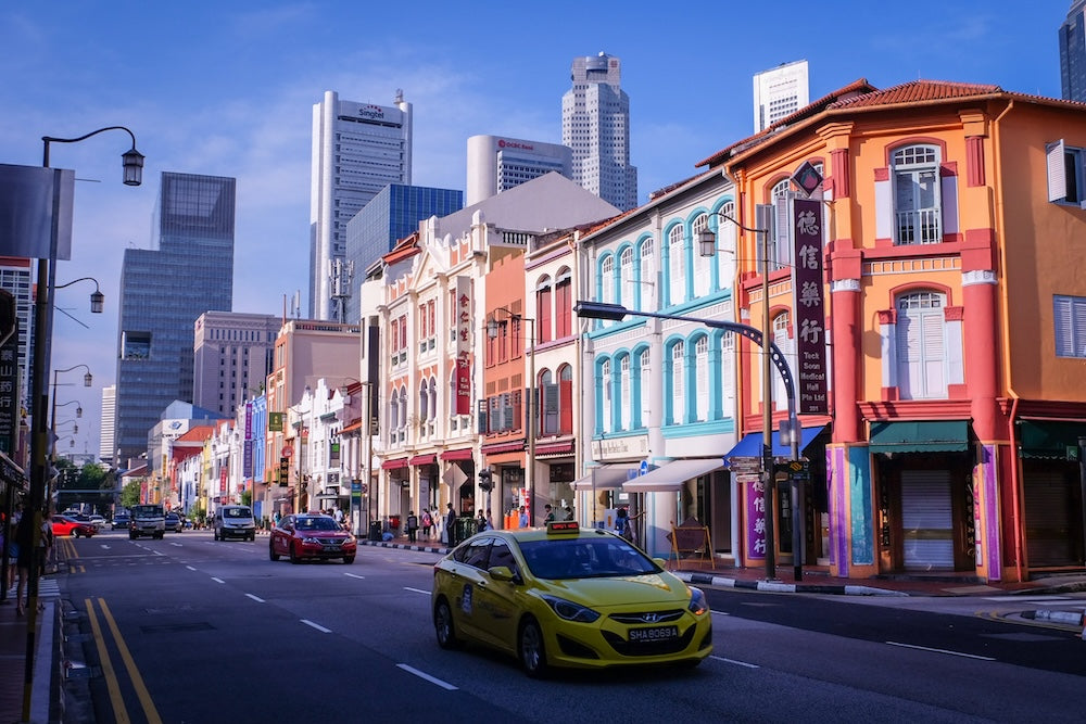 A yellow Comfort taxi driving in front of a red SMRT cab along colourful shophouses on South Bridge Road, with skyscrapers, including the Singtel building, in the background on a sunny day in Singapore.