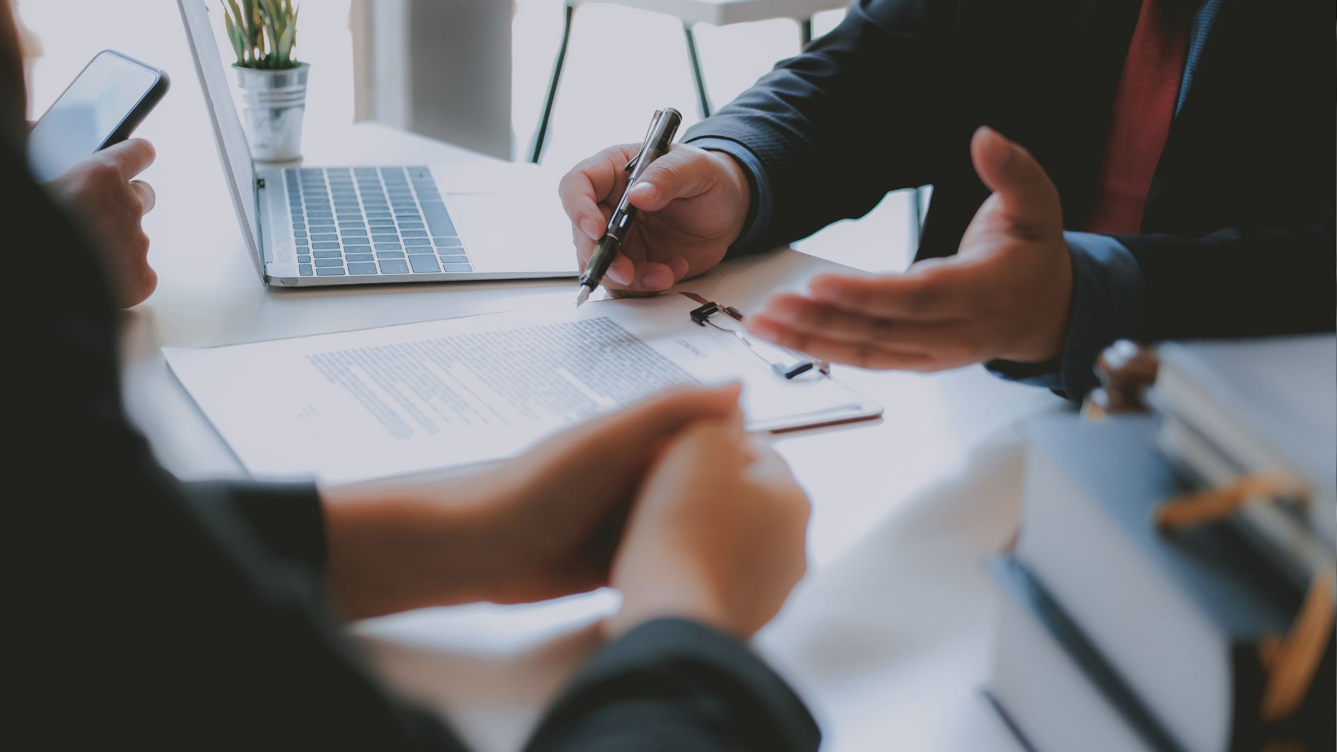Close-up of a business meeting with two people discussing a contract, one holding a pen and gesturing, with a laptop and documents on the table.
