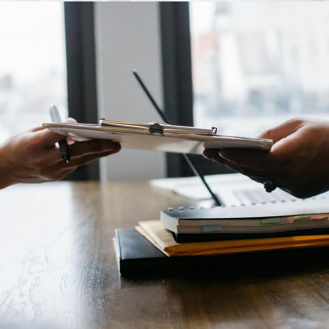 Two people exchanging a clipboard over a desk with notebooks, a laptop, and papers in an office setting.