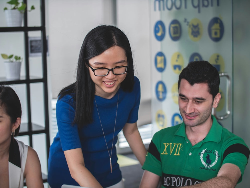 Woman in blue helping a man in green at a desk, both smiling in a bright office setting.