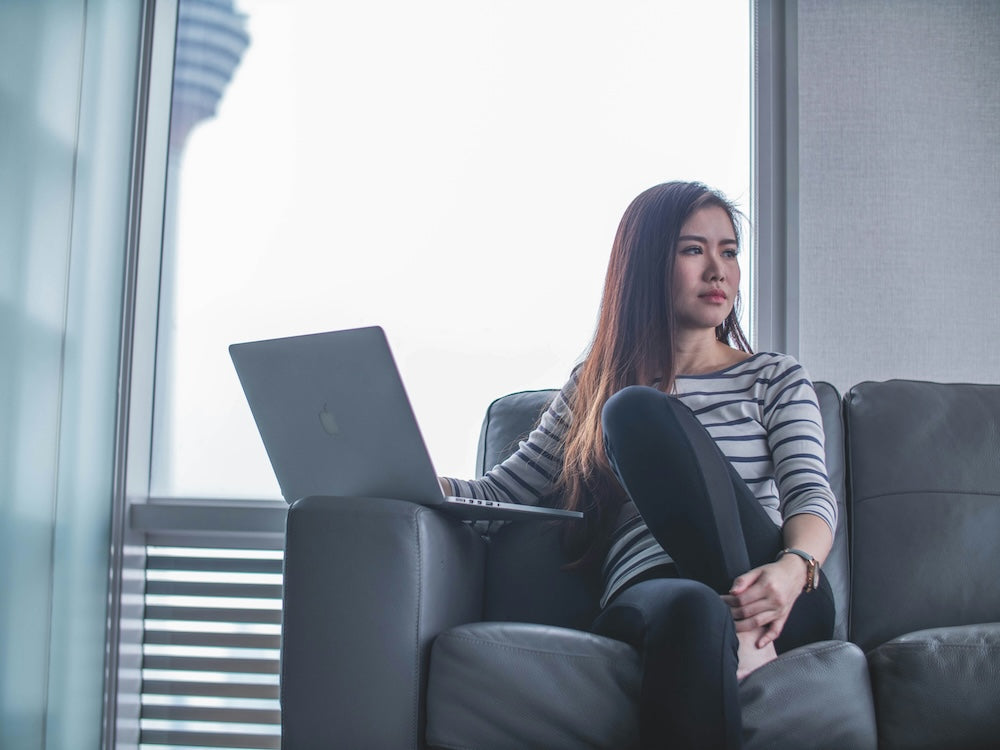 Woman sitting on a sofa with a laptop, looking thoughtfully out the window in a bright modern room.