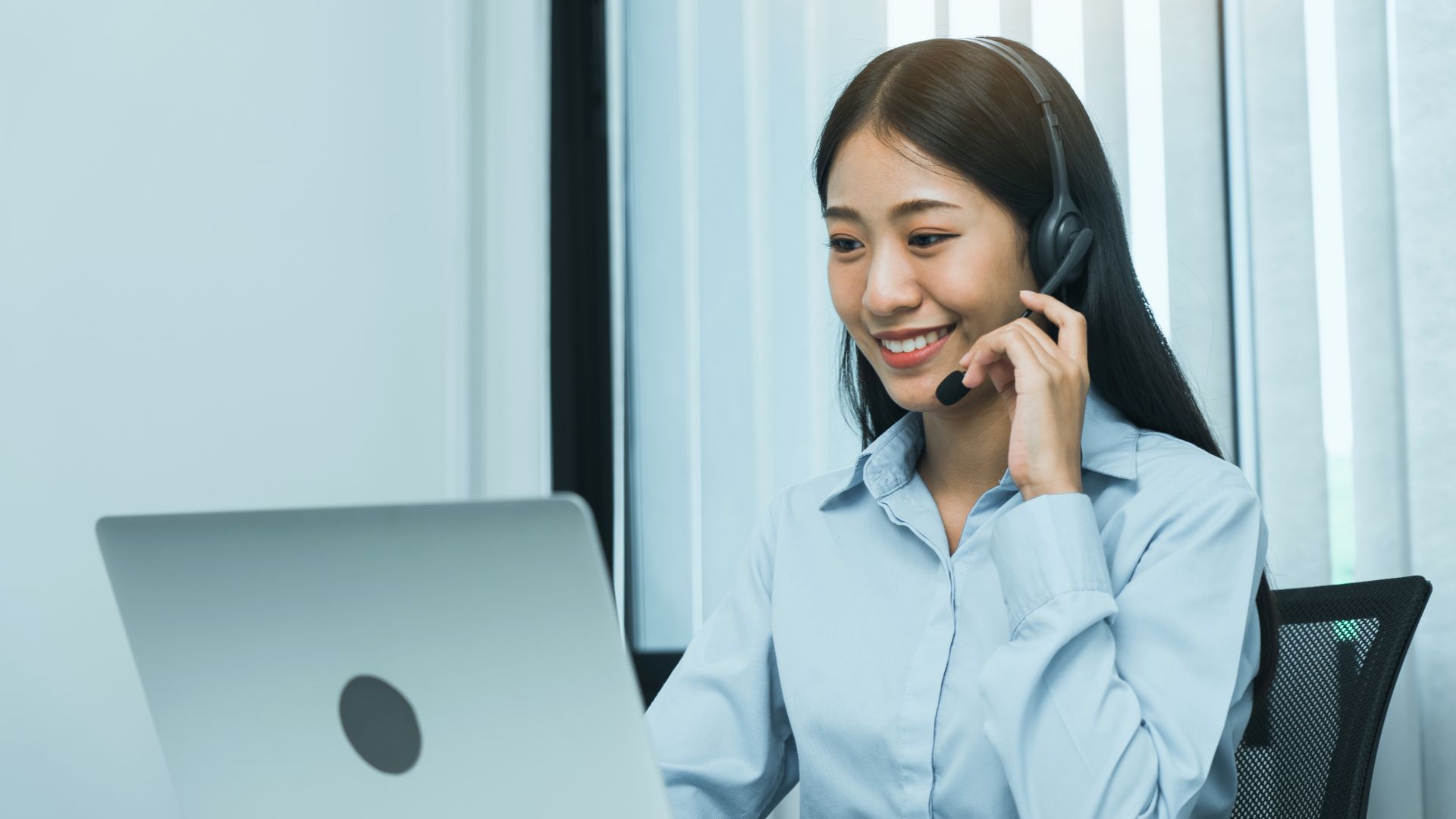 Smiling woman wearing a headset, working on a laptop in a bright office with blinds in the background.