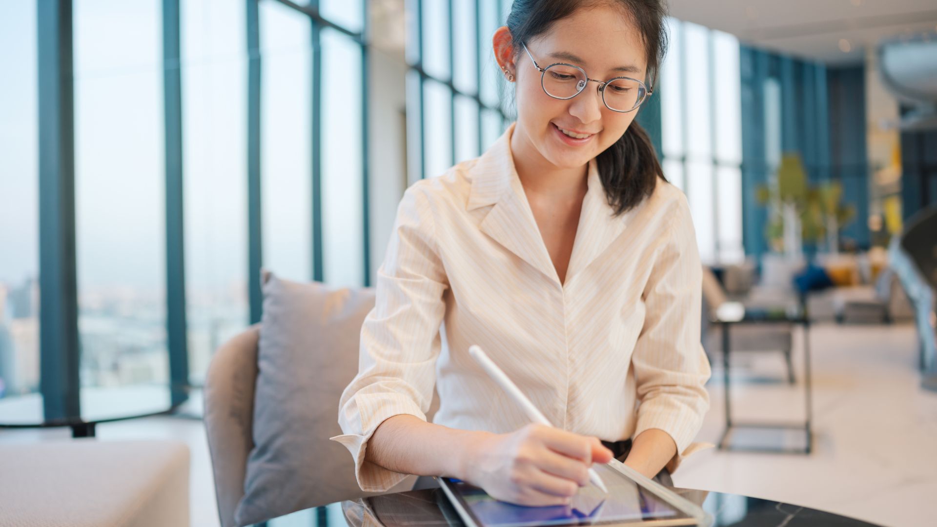 Smiling woman using a digital tablet with a stylus in a bright, modern office lounge with large windows.