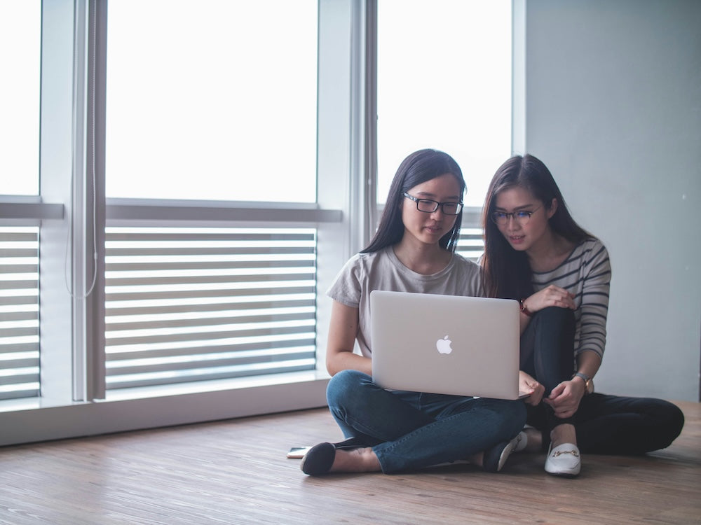 Two women sitting on the floor, working together on a laptop in a bright room with large windows.