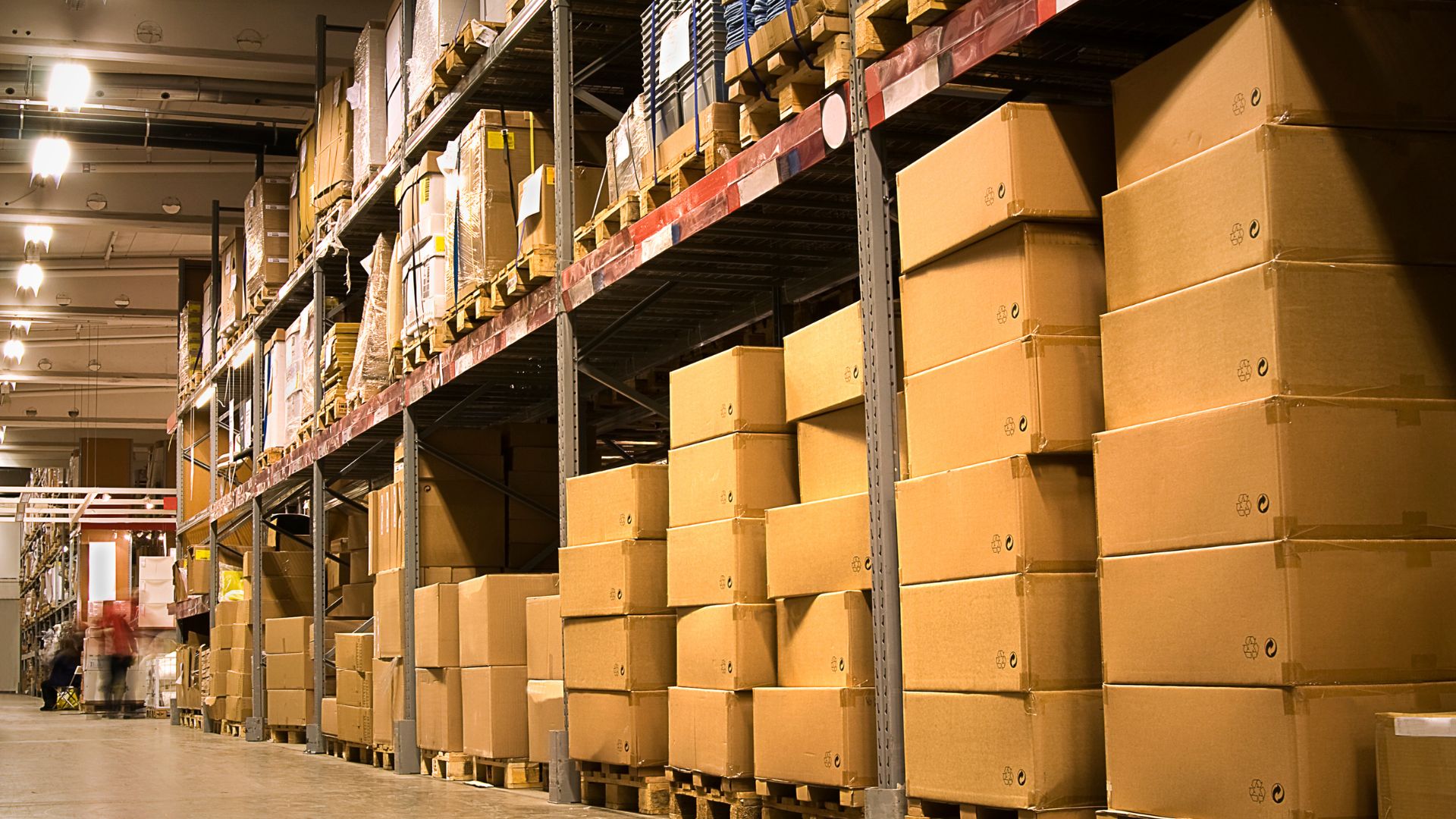 Warehouse interior with shelves stacked with cardboard boxes and pallets, organised for storage and distribution.