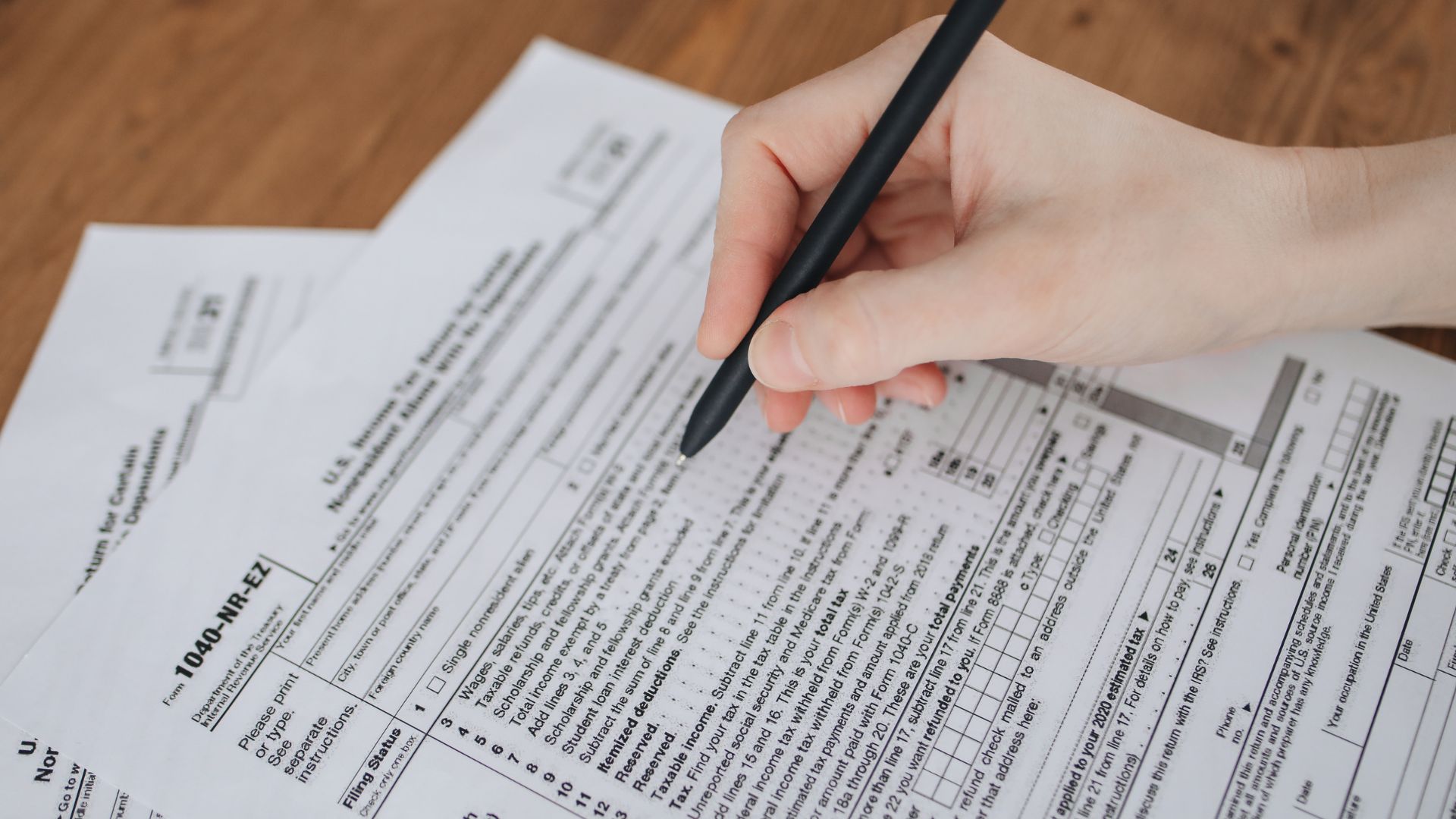 Close-up of a hand filling out official forms with a pen on a wooden desk, completing detailed paperwork and documents.