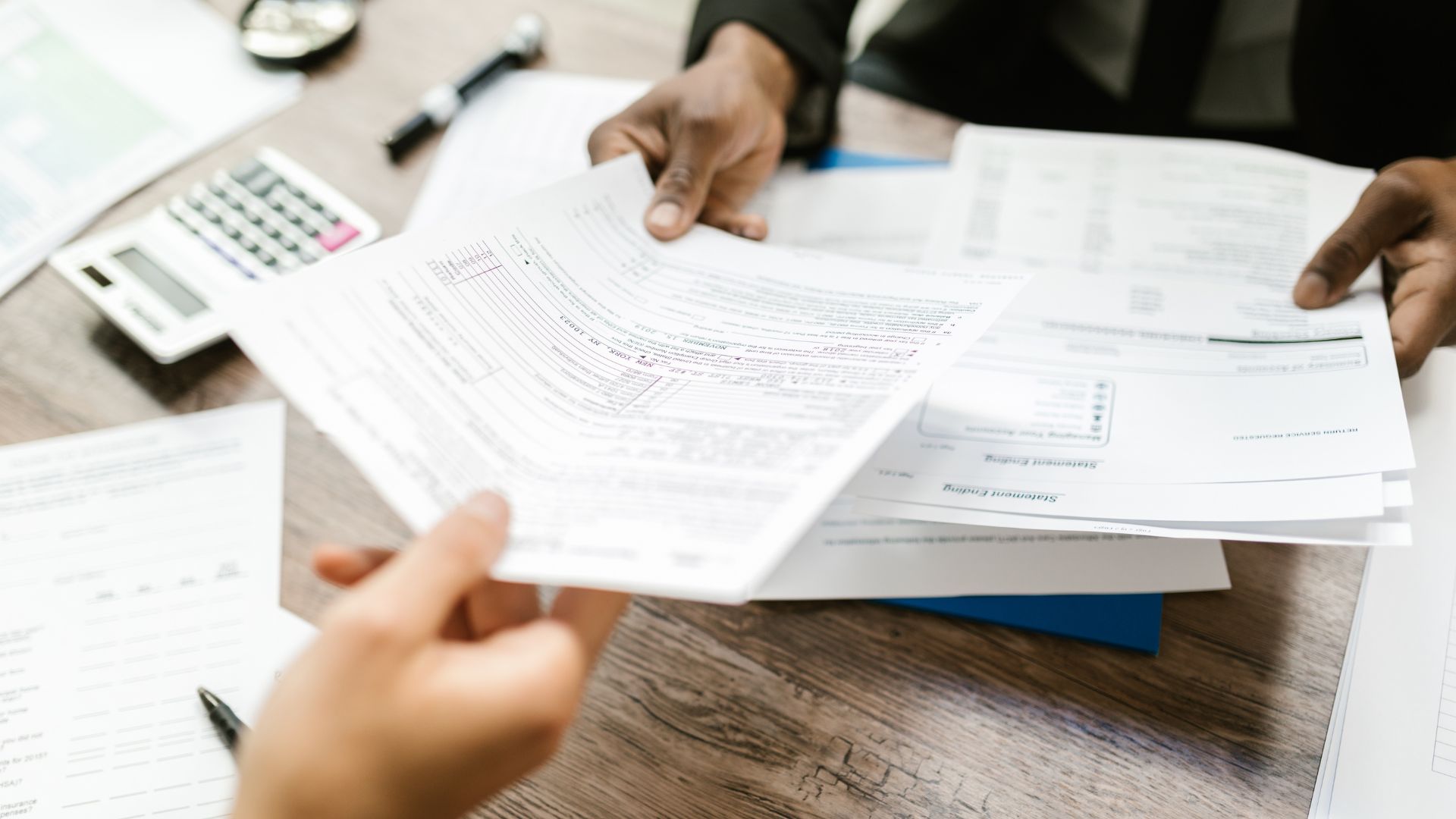 Two people exchanging and reviewing paperwork at a desk with documents, a calculator, and a pen.