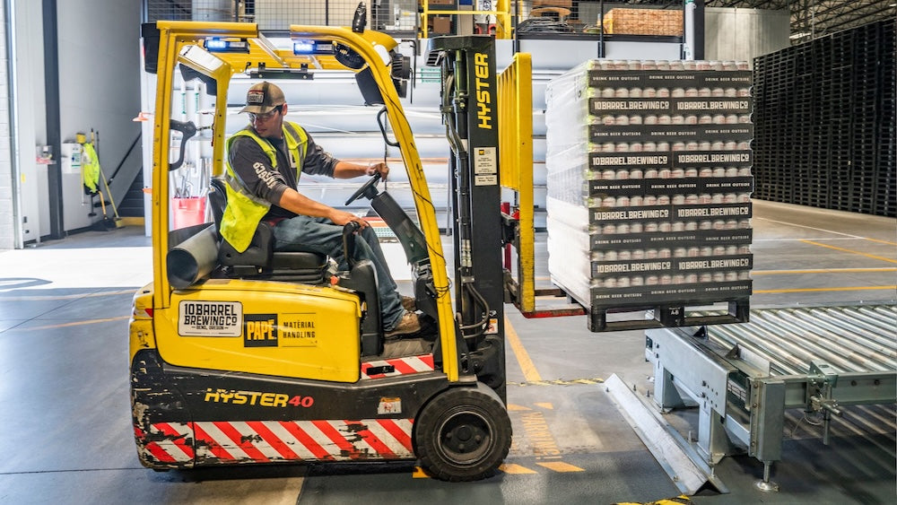 Warehouse worker driving a yellow forklift, moving a pallet of boxes onto a conveyor inside an organised warehouse facility.