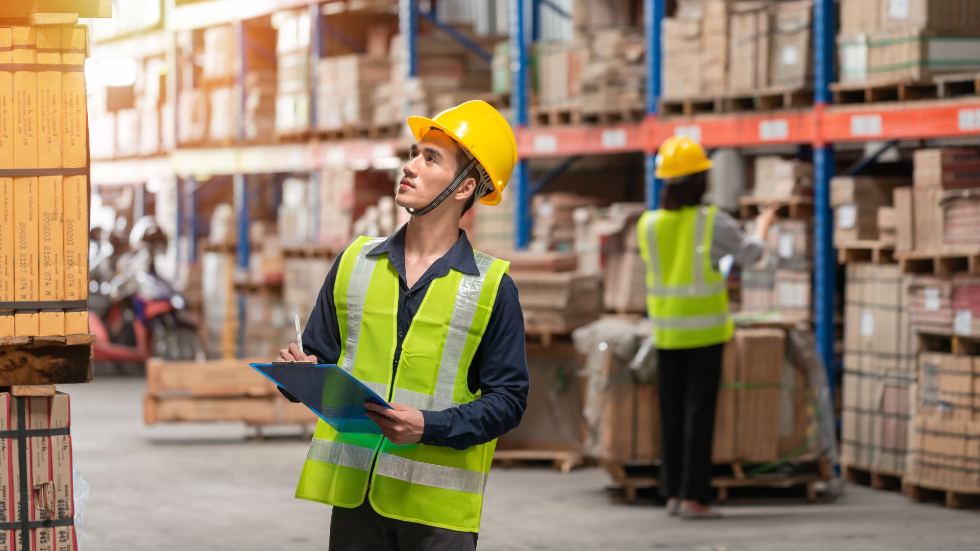 Warehouse worker in safety gear holding a clipboard and inspecting shelves while another worker organises boxes in the background.