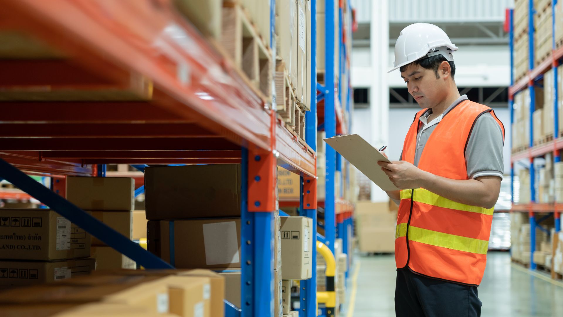 Warehouse worker wearing safety gear checking inventory on shelves and writing notes on a clipboard inside a storage facility.