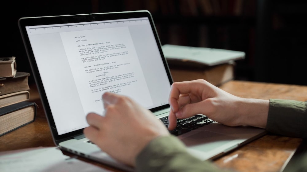 Person typing on a laptop displaying a document, with books and papers on the wooden desk.