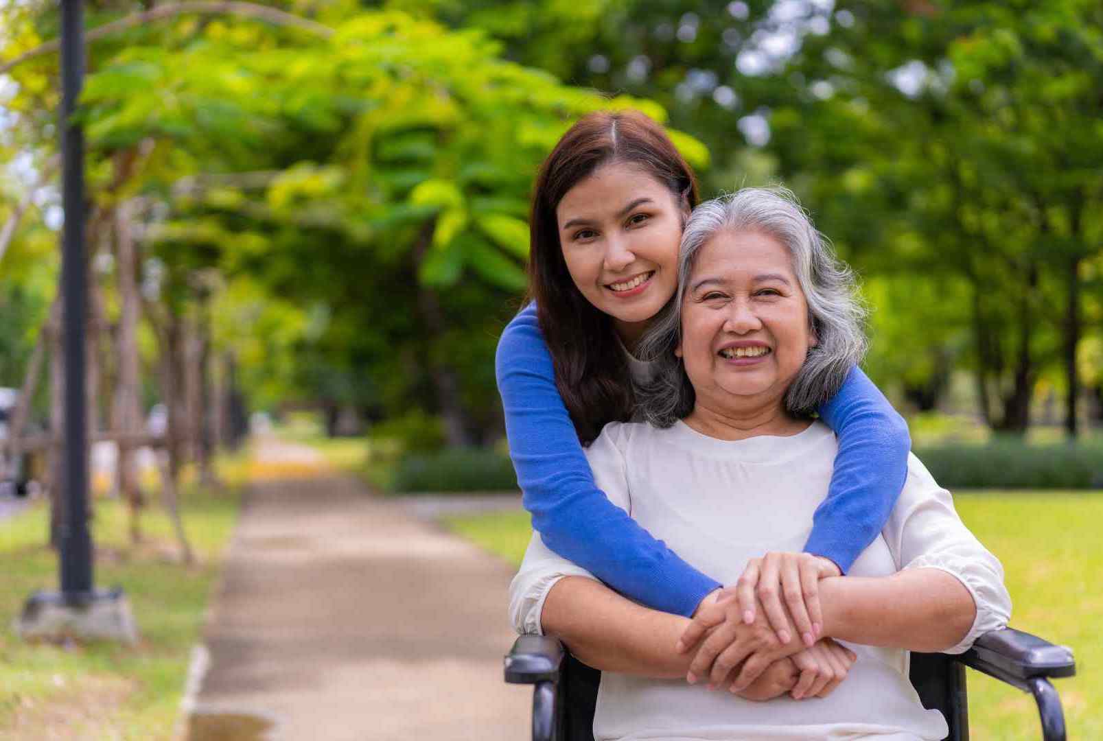Elderly woman in wheelchair smiling with younger woman hugging her from behind in a park, showing warmth and elderly care support.