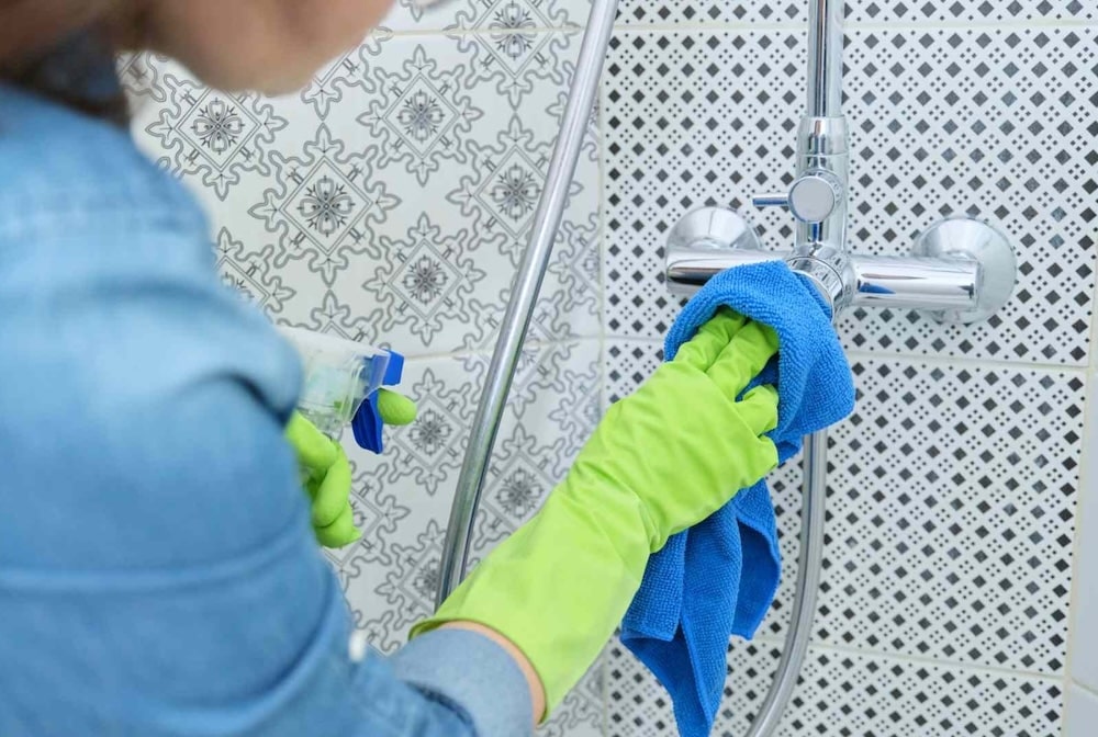 Person wearing green gloves cleaning a bathroom faucet with a blue cloth and spray bottle against patterned tiles.