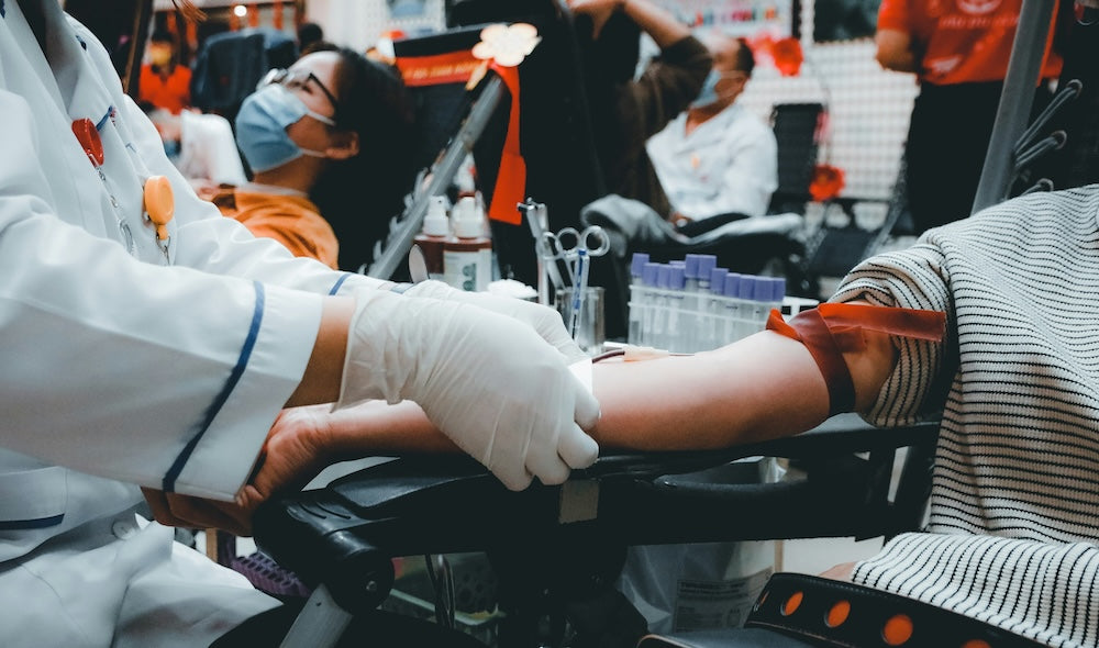 "A gloved doctor in a lab coat applying a bandage after drawing blood, with a masked middle-aged woman wearing spectacles in the background.