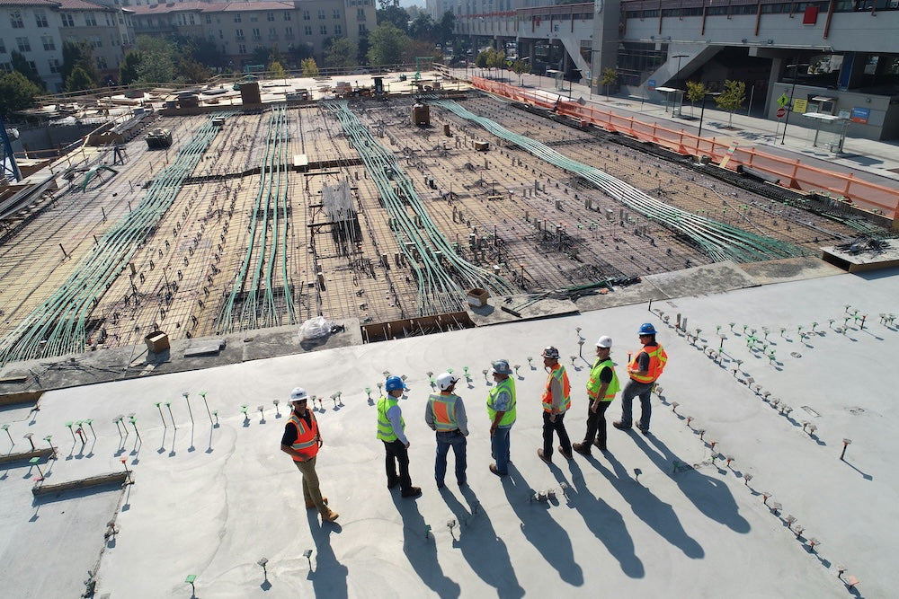 Workers in safety gear standing on a large construction site inspecting concrete foundation work under bright sunlight
