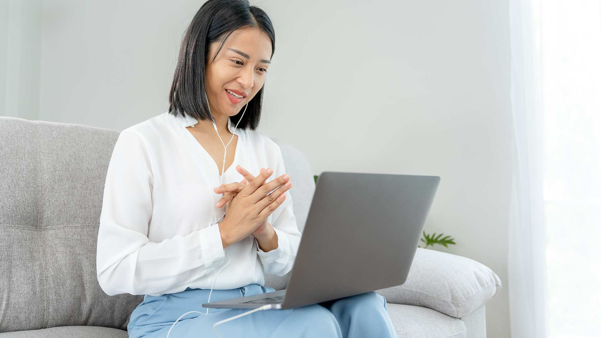 Smiling woman sitting on a couch, wearing earphones and talking during a video call on her laptop.